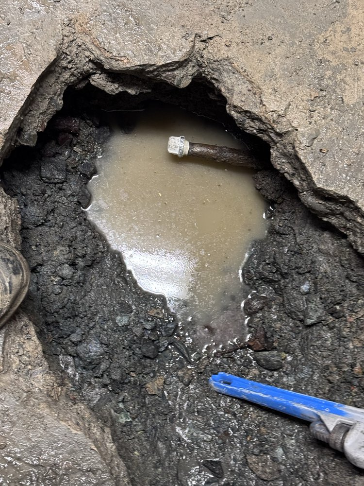 Close-up of a corroded underground pipe fitting leaking water, exposed during an emergency repair